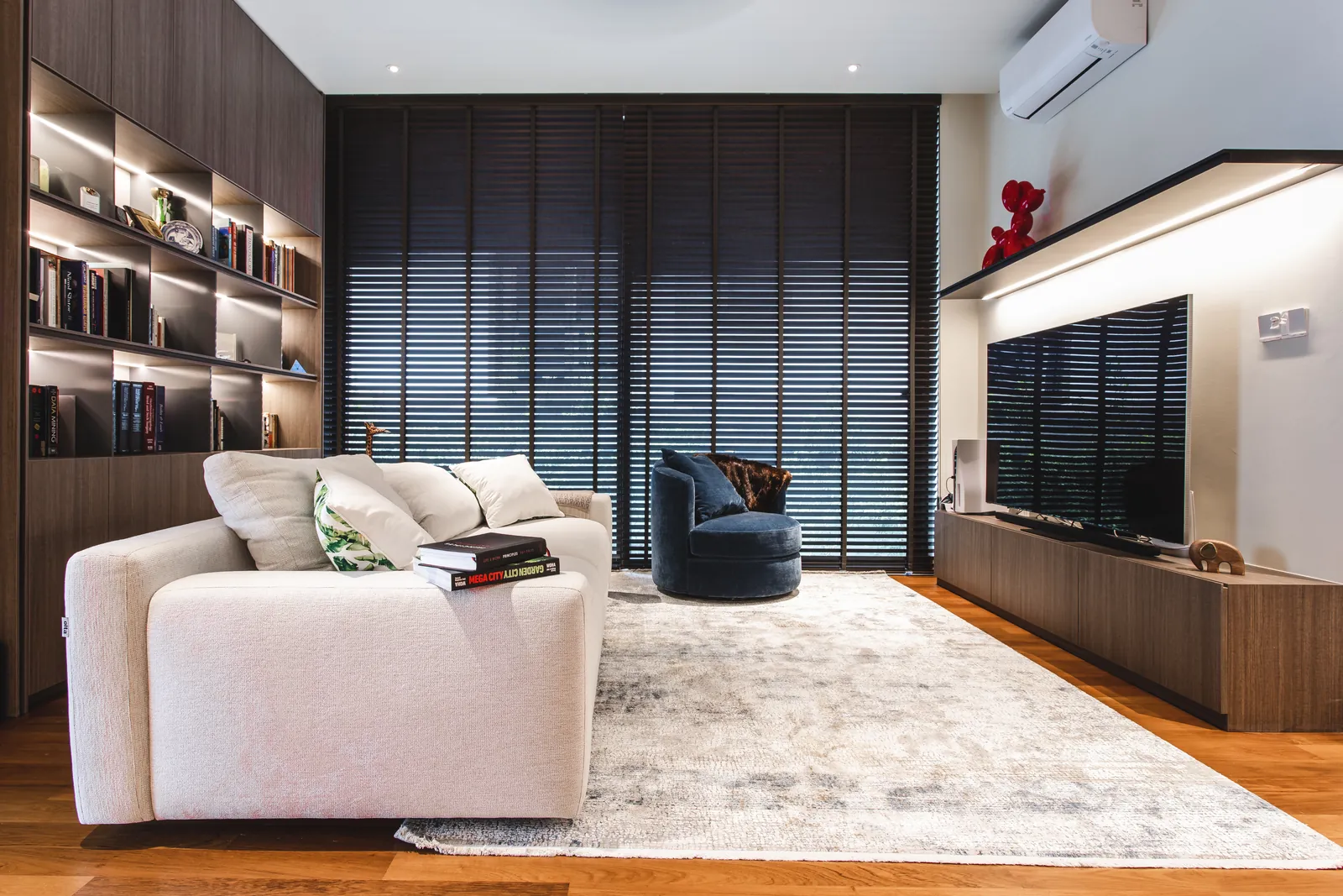 House of the Collector: family room with a cream sofa, a velvet armchair, lit bookshelves and timber blinds