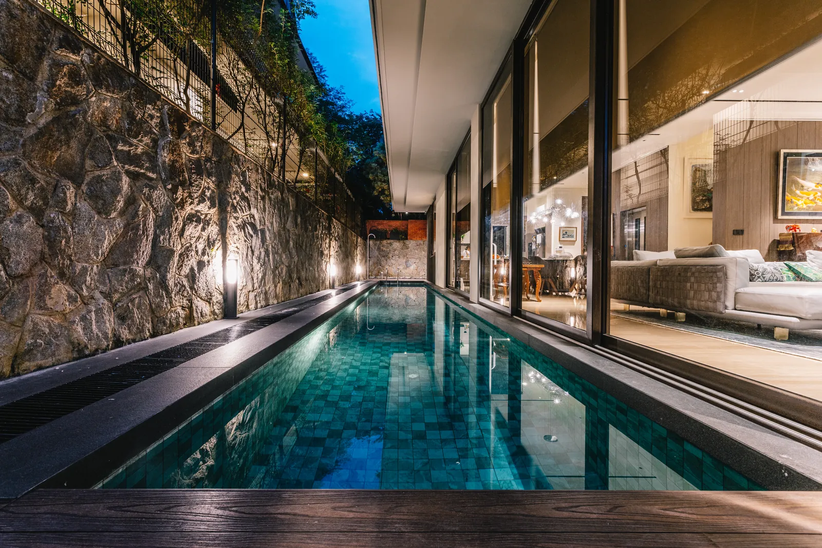 House of the Collector: lap pool along a lit rubble-stone wall at dusk, beside the living room