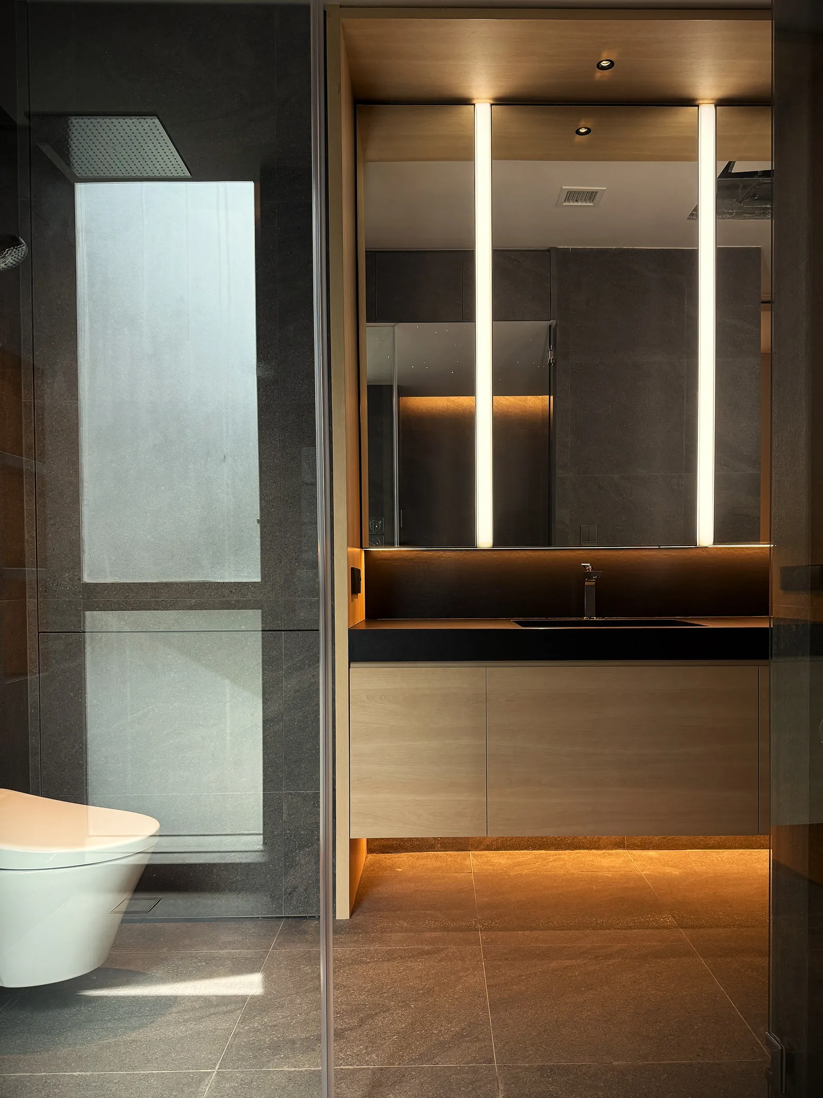 The Garden Within: bathroom in dark stone and timber, with lit mirror strips and a frosted window